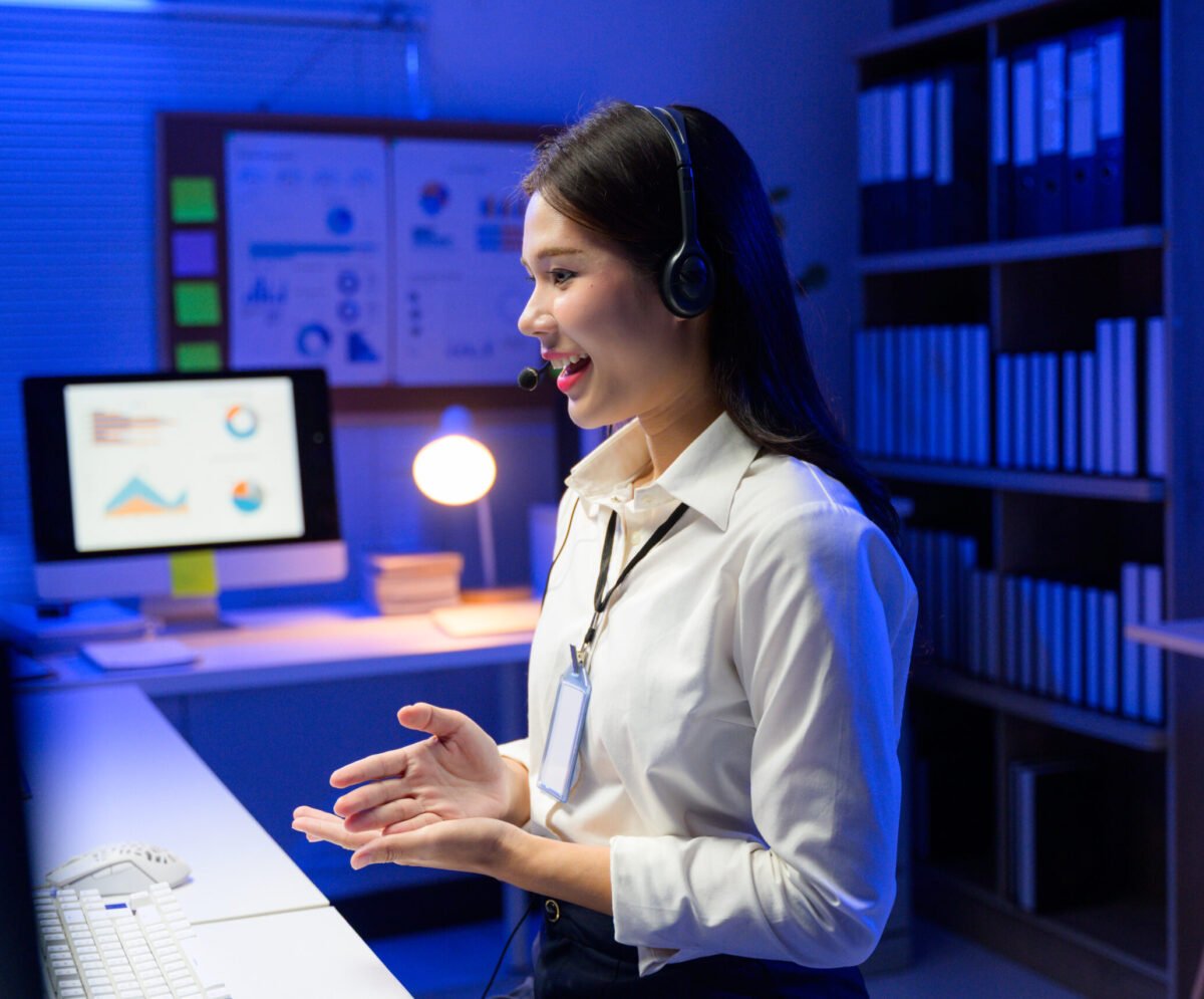 Asian businesswoman working late using headset in office with computer showing graphs